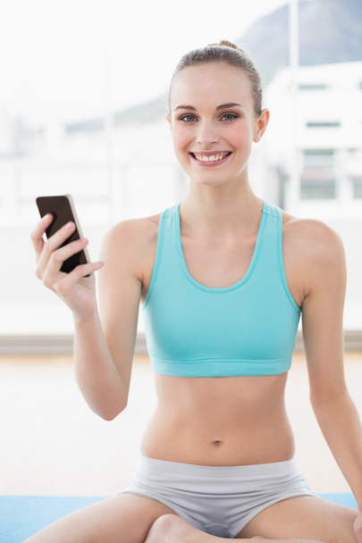 Deportiva mujer sonriente sosteniendo el teléfono mientras está sentado en la esterilla de ejercicio
 - Foto, imagen