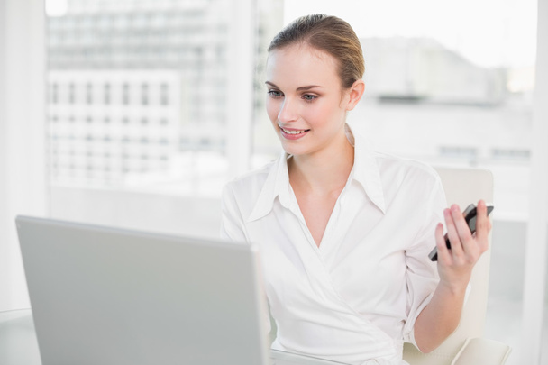 Cheerful businesswoman using laptop and texting on smartphone - Photo, image