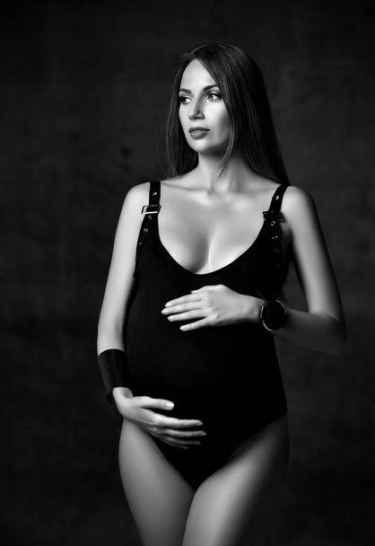 Young beautiful slim pregnant woman with long hair in black body and accessories standing, embracing belly and looking aside - Foto, Imagem