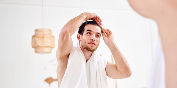 Νεαρός αξύριστος άντρας χτενίζει τα μαλλιά του δίπλα στον καθρέφτη στο μπάνιο. - Φωτογραφία, εικόνα
