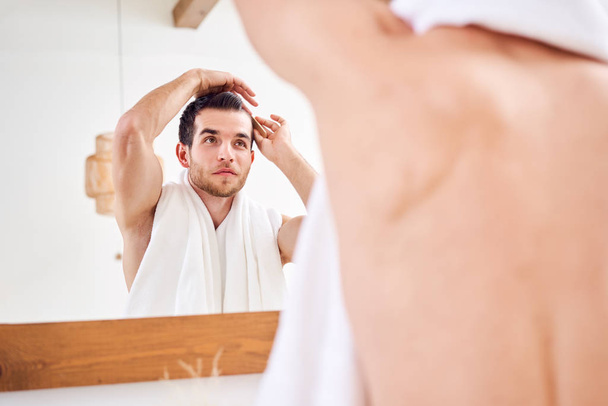Man combing his hair while standing with white towel on neck near mirror in bathroom - Foto, Imagem