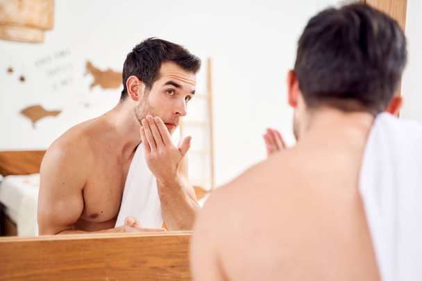 Sportive man lathers face and stands with towel on his shoulders in front of bathroom mirror - Fotografie, Obrázek