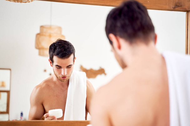 Young sporty man with towel on his shoulders with jar of cream in his hands stands in front of bathroom mirror - Foto, imagen