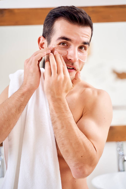 Male applies transparent patches under his eyes while standing in bath opposite mirror - Φωτογραφία, εικόνα