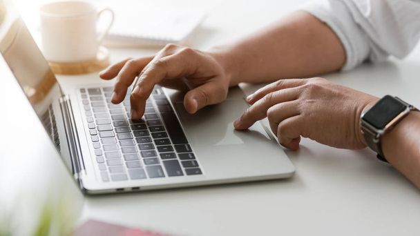 Vista lateral do empresário digitando no laptop em espaço de trabalho simples com xícara de café e material de escritório
 - Foto, Imagem