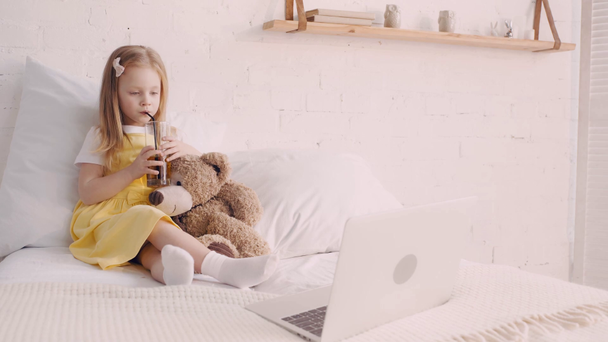 Yatakta dizüstü bilgisayarda film izlerken oyuncak ayıdan meyve suyu içen çocuk. - Video, Çekim