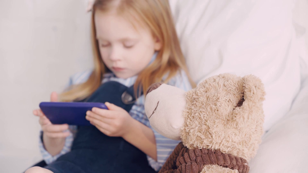 Yatakta oyuncak ayının akıllı telefonunu kullanan seçici bir çocuk. - Video, Çekim
