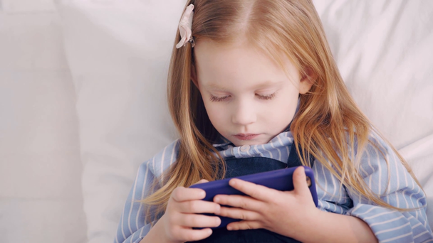 Cute kid za pomocą smartfona na łóżku - Materiał filmowy, wideo