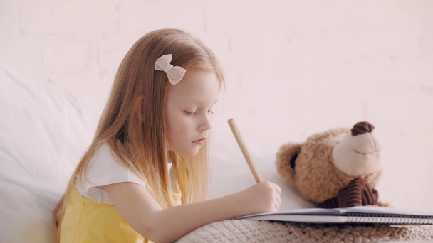 Cute kid drawing on bed by teddy bear - Footage, Video