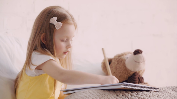 Child drawing with pencil by teddy bear on bed - Footage, Video