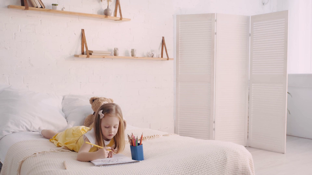 Cute child drawing on bed by teddy bear - Footage, Video