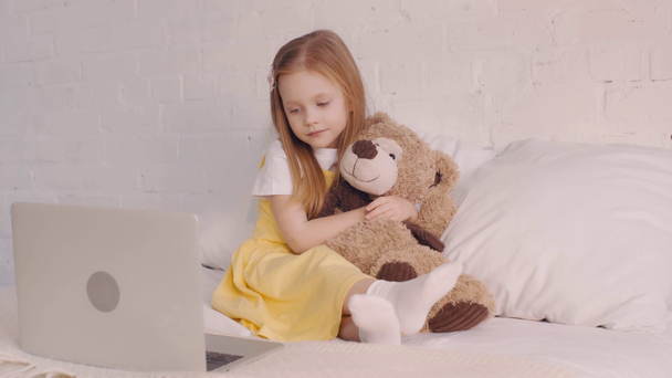 Kid przytulając misia podczas oglądania filmu na laptopie w łóżku - Materiał filmowy, wideo