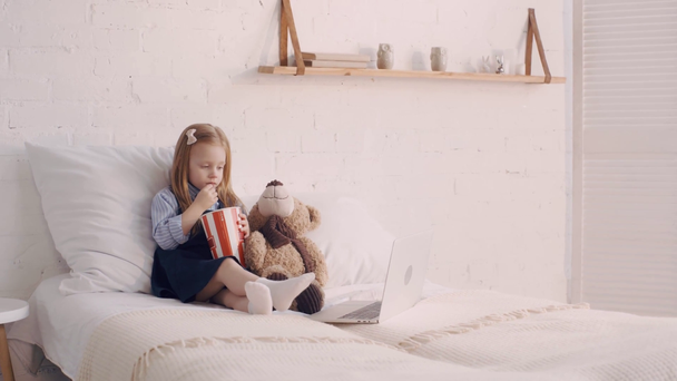 Dítě jíst popcorn při sledování filmu na notebooku plyšového medvídka na posteli - Záběry, video