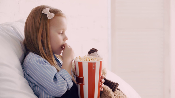 Criança comendo pipoca por ursinho de pelúcia e assistindo desenhos animados no laptop no quarto
 - Filmagem, Vídeo