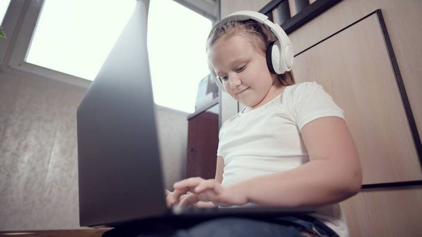A smart little girl of seven years old in white headphones with a laptop in her hands is pushing on the floor in her room. The young generation on the Internet and IT technology - Фото, зображення