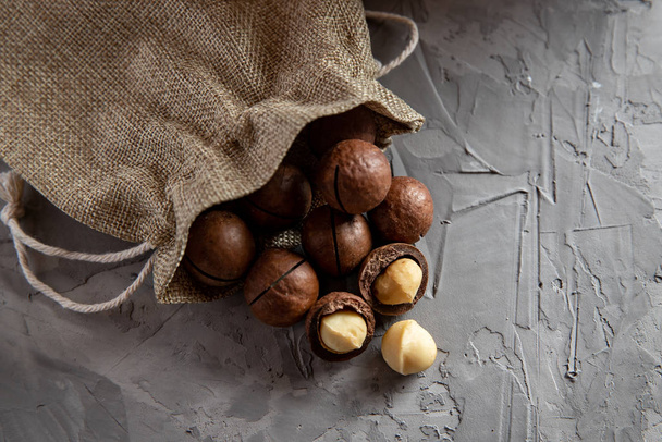 Pile macadamia nuts open kernels and shells in burlap bag on gray concrete background, top view - Фото, изображение