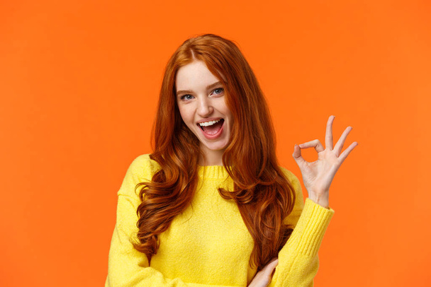 Sassy and daring cool redhead girl with curls, showing okay excellent gesture and smiling excited, assure party be awesome, give permission, say yes, confirm or recommend something, orange background - Photo, Image