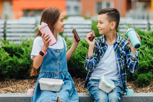 boy and girl sitting near building with chocolate cakes and thermoses in hands  - Foto, immagini