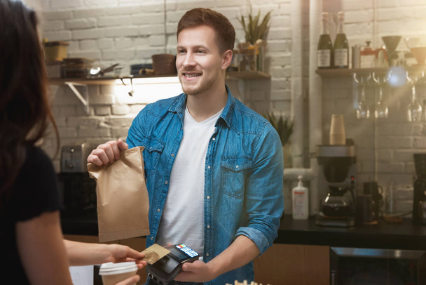 hospitable barista man standing behind bar sells hot coffee drink and lunch in paper bag to woman client for takeaway , woman pays with bank card - Photo, image