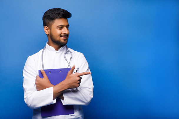 Portrait of friendly smiling male doctor with clipboard on blue background - Foto, imagen