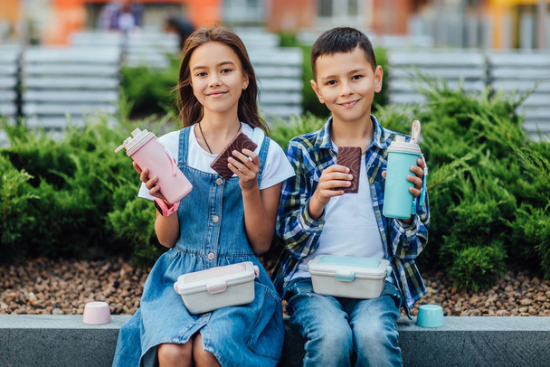 boy and girl sitting near building with chocolate cakes and thermoses in hands  - Fotó, kép