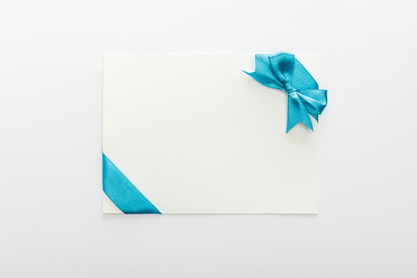 top view of blank card with blue decorative satin ribbon and bow on white background - Fotó, kép