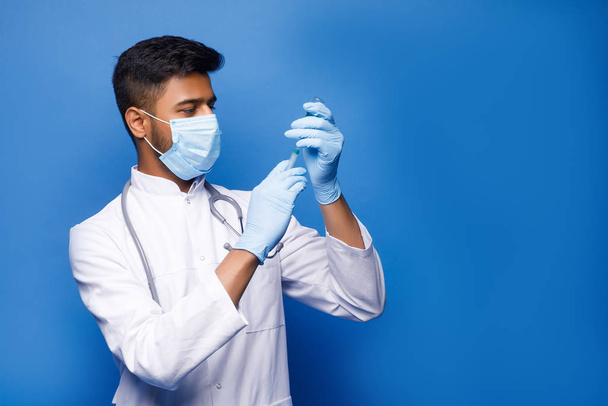 Focused indian doctor male looking at syringe with medicine on blue studio background - Photo, Image