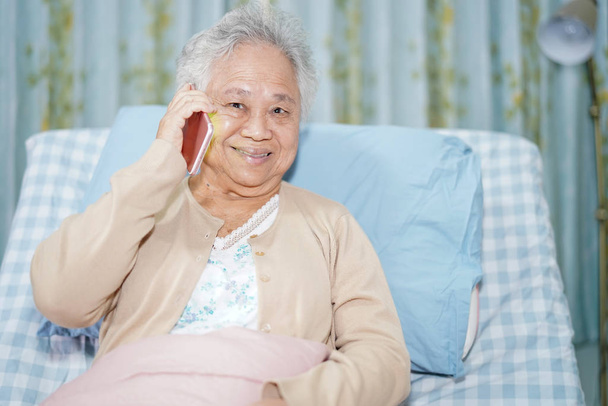 Azjatycka starsza lub starsza pani pacjentka rozmawiająca przez telefon komórkowy, siedząca i szczęśliwa w łóżku na oddziale pielęgniarskim: zdrowa, silna koncepcja medyczna - Zdjęcie, obraz