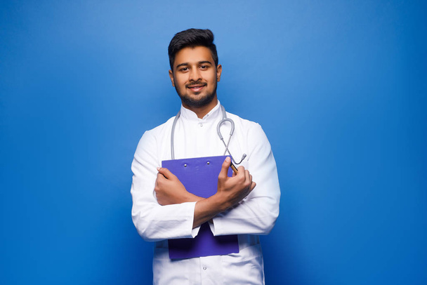 Portrait of friendly smiling male doctor with clipboard standing on blue background - Foto, Bild