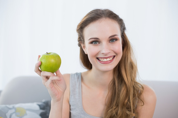 Happy young woman sitting on sofa holding green apple - Photo, Image