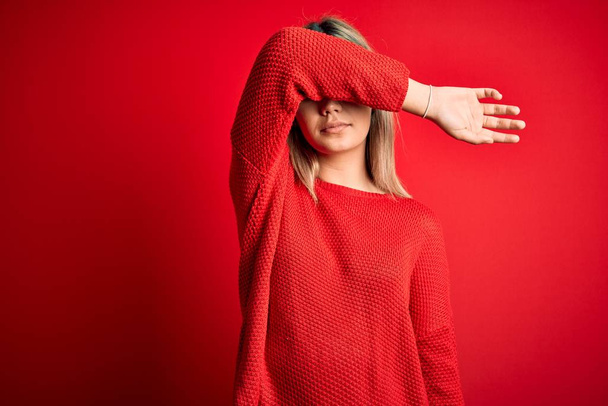 若い美しいブロンドの女性は、腕で目を覆う赤い隔離された背景にカジュアルなセーターを着て、真剣に悲しい顔をしています。見えない、隠れている、拒絶の概念 - 写真・画像