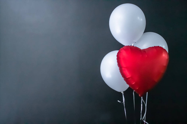 Red heart-shaped balloon and white round air balloons on a black background. Valentines Day, holiday, love. - 写真・画像