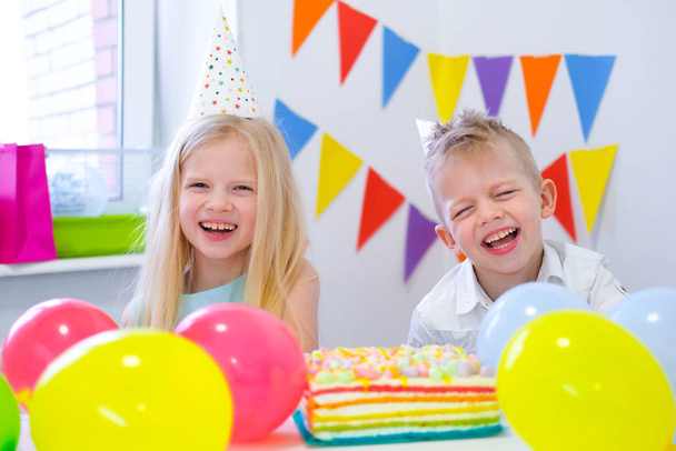 Dva blonďatí běloši kluci a holka se baví a smějí se na narozeninové oslavě. Barevné pozadí s balónky a narozeninový duhový dort. - Fotografie, Obrázek