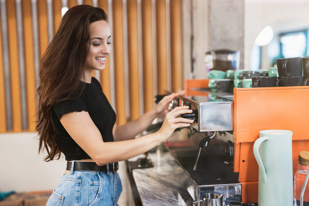 young smiling barista woman working behind the bar, stands near professional coffee machine - Fotografie, Obrázek