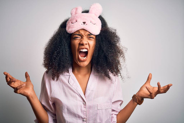 Mujer afroamericana joven con cabello afro usando máscara para dormir y pijama rosa loca y loca gritando y gritando con expresión agresiva y brazos levantados. Concepto de frustración
. - Foto, Imagen
