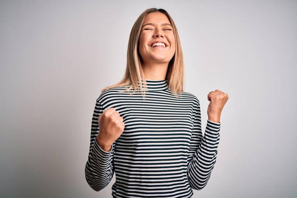 Young beautiful blonde woman wearing casual striped sweater over isolated background very happy and excited doing winner gesture with arms raised, smiling and screaming for success. Celebration concept. - Photo, Image