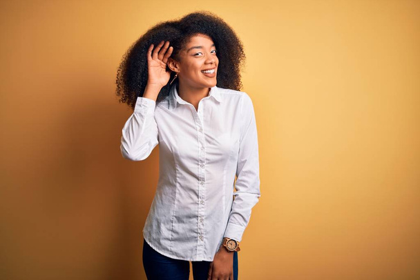 Fiatal, gyönyörű afro-amerikai elegáns nő afro hajjal áll sárga háttér felett mosolyogva, fültől fülig hallgatva egy pletykát vagy pletykát. Süketség fogalma. - Fotó, kép