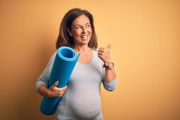 Middle age pregnant woman expecting baby holding yoga mat for healthy lifestyle pointing and showing with thumb up to the side with happy face smiling - Fotografie, Obrázek
