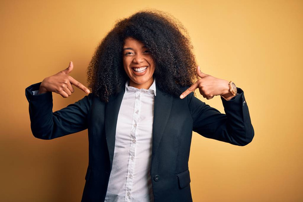 Young beautiful african american business woman with afro hair wearing elegant jacket looking confident with smile on face, pointing oneself with fingers proud and happy. - 写真・画像