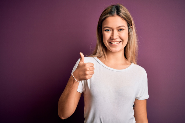 Young beautiful blonde woman wearing casual white t-shirt over purple isolated background doing happy thumbs up gesture with hand. Approving expression looking at the camera showing success. - 写真・画像