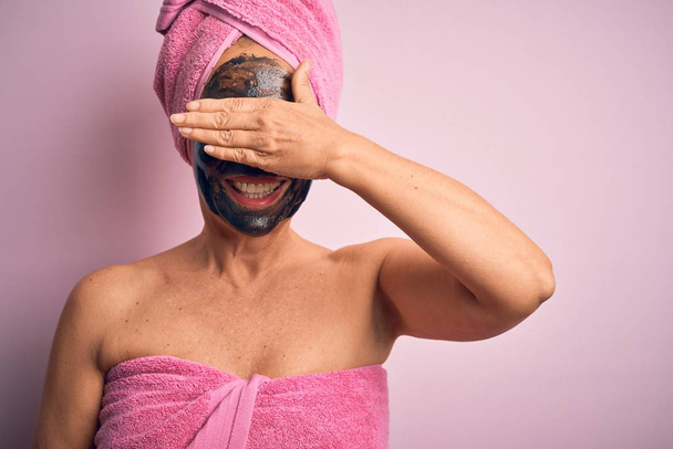 Middle age brunette woman wearing beauty black face mask over isolated pink background smiling and laughing with hand on face covering eyes for surprise. Blind concept. - Foto, immagini