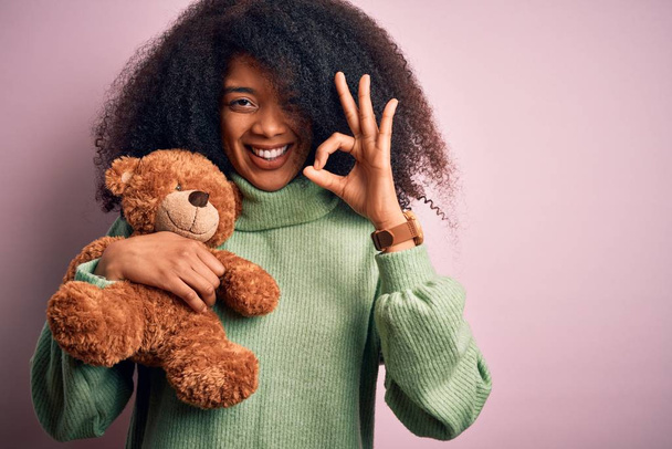 Young african american woman with afro hair hugging teddy bear over pink background doing ok sign with fingers, excellent symbol - Фото, изображение
