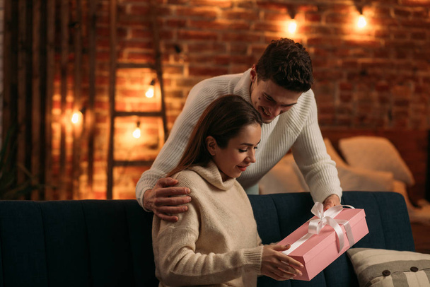 Handsome man presenting a gift to his beautiful girlfriend and smiling. Beautiful young couple at home enjoying spending time together.Valentines Day, relationships and people concept - Foto, afbeelding