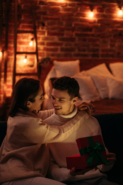 Handsome man presenting a gift to his beautiful girlfriend and smiling. Beautiful young couple at home enjoying spending time together.Valentines Day, relationships and people concept - Zdjęcie, obraz