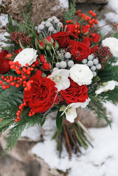 In winter, a brides bouquet of red and white flowers ranunculus, spray rose, brunia, ilex, hellebore, and burgundy skimmy stands in a transparent vase in the snow in a vase. Vertical large frame. - Foto, Bild