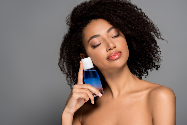 nude african american girl with closed eyes holding bottle of makeup remover, isolated on grey - Foto, immagini