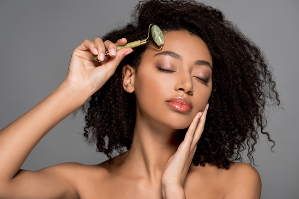 beautiful african american girl with closed eyes using jade roller, isolated on grey - Fotó, kép