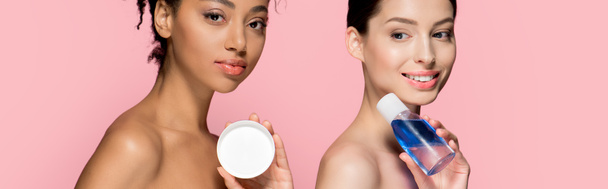 panoramatický záběr atraktivní multietnické dívky držící kosmetický krém a odstraňovač make-upu, izolované na růžové - Fotografie, Obrázek