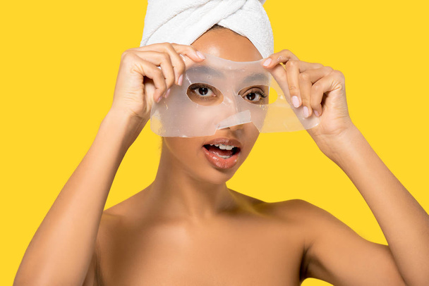 portrait of shocked african american girl with towel on head applying hydrogel mask, isolated on yellow - Photo, Image