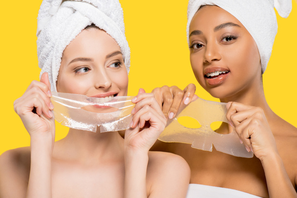 portrait of multicultural women with towels on heads holding hydrogel masks, isolated on yellow - Foto, imagen